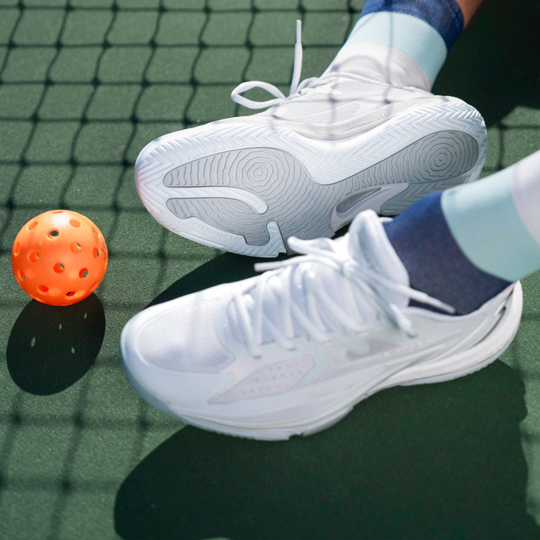 A pair of white athletic sneakers worn with white socks and purple cuffs, shown on a tennis court net with a tennis ball nearby.